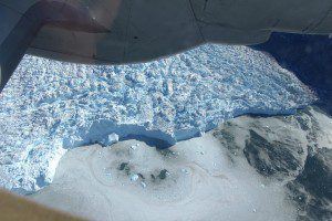 A top-down view from a NOAA P-3 aircraft shows the calving front of the Sermeq Kujatdleq glacier. The aircraft's lower engine nacelle and left main landing gear fairing is in the foreground at the top. (Credit: NASA/John Sonntag)